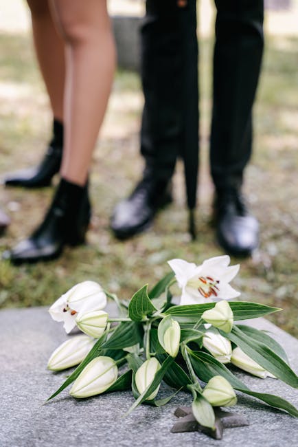 A close-up of a floral arrangement placed on the ground, featuring white lilies with open blooms revealing their orange and brown stamens, surrounded by several white buds and long, slender green leav