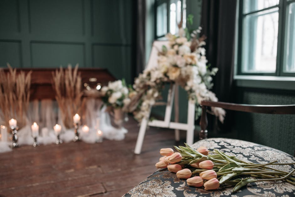 A floral arrangement scene featuring a bouquet of pale pink tulips with green stems resting on an ornate upholstered chair in the foreground. In the background, there is a blurred display of white flo