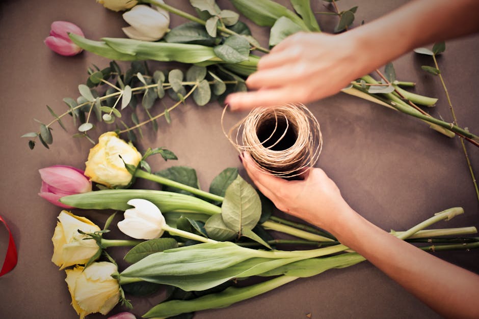 A person's hands are arranging a floral bouquet on a dark, matte surface, with a spool of natural twine held in one hand and the other hand adjusting a stem. The bouquet includes fresh, pastel-colored