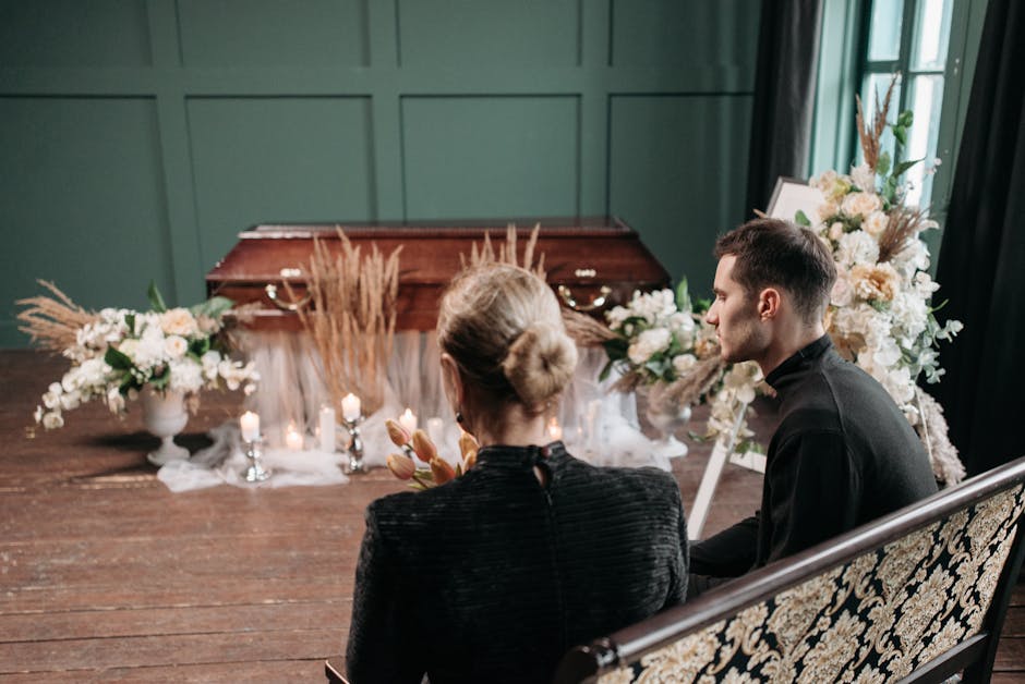 The image depicts two individuals seated side by side on an ornate, vintage-style bench in a softly lit room, facing a memorial display. They are dressed in dark clothing appropriate for a funeral set