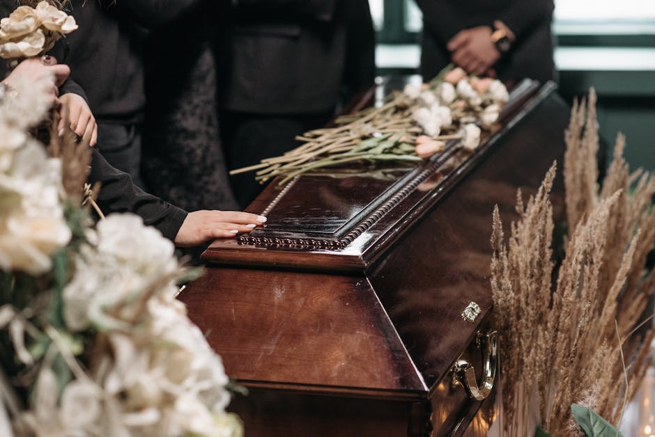 A close-up of a polished dark wooden casket with a glossy finish, adorned with an arrangement of light pink and white flowers, including roses and lilies, resting on top. Surrounding the casket are ta