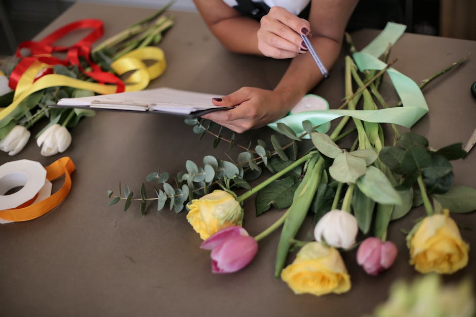 A florist is seated at a table, meticulously preparing a floral arrangement for flower delivery, as part of a floral care checklist for event planners. The workspace features an assortment of freshly 