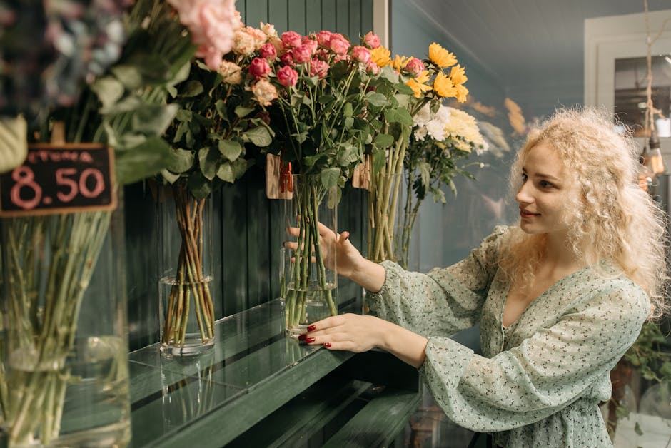 A woman with blonde, curly hair and fair skin choosing fresh flowers in a florist shop. She is wearing a light, patterned blouse with puffed sleeves and has red nail polish. She is reaching out with h