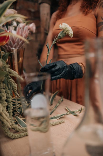 A person wearing a brown top and black gloves is holding a white flower with a long green stem in their right hand during a floral arrangement demonstration at a workshop. The workspace features a woo
