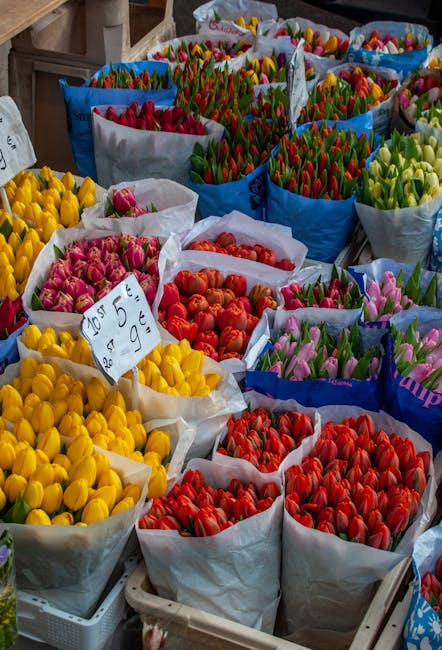 A vibrant display of tulip bouquets arranged in colorful paper wrapping at a flower market, showcasing a variety of shades including yellow, pink, red, and green. The tulips appear fresh with smooth p